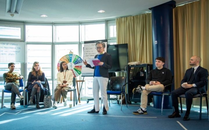 In a conference room, a presenter stands by a whiteboard and colorful wheel, addressing a seated audience.