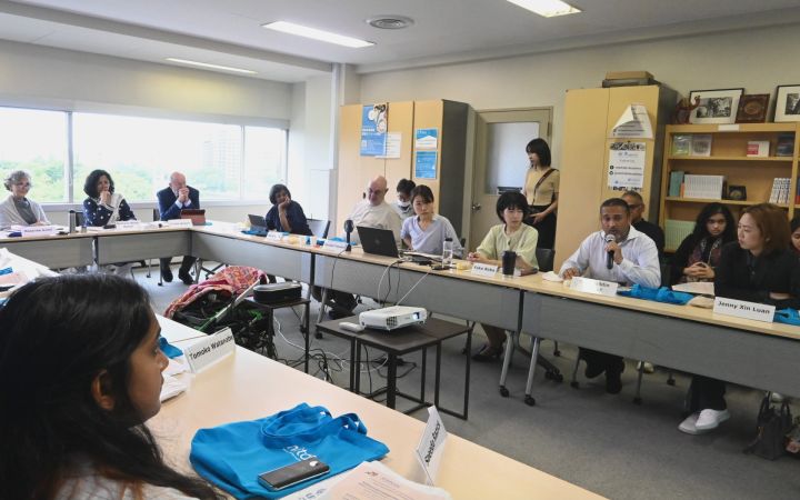 A group of people in a roundtable discussion. On the lower left side of the frame is a woman listening to a man who is speaking across the table while sitting with other meeting participants. Placed on the table in front of the woman is a blue tote bag bearing the UNITAR logo (not fully visible), a smartphone, and her nameplate.