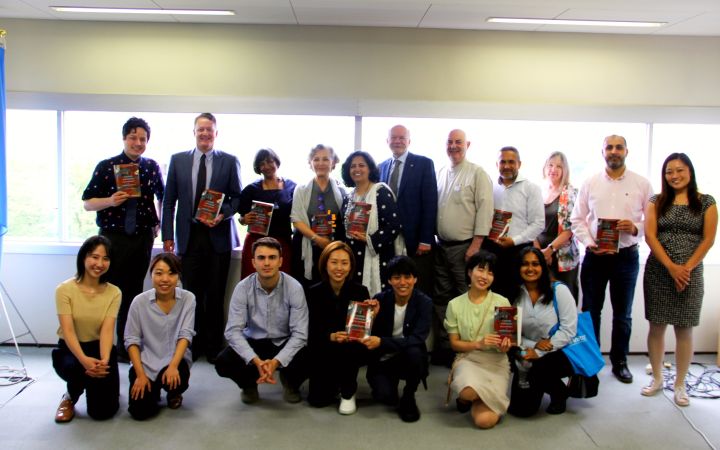 A group of people posing for a photo with glass window panels as main background. The back row consists of men and women standing where mostly are holding a book. At the front row are men and women kneeling with some also holding a book. On the leftmost side of the photo is a UN flag (not fully visible) hanging on a standee.