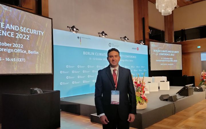 Dr. Sameh Al Muqdadi smiling with arms crossed, behind him are the flags of various countries at the UN Headquarters