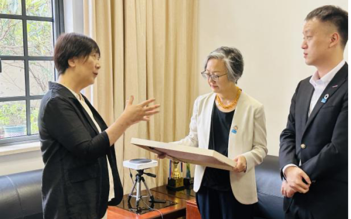 In an office with a bright window, an Asian woman in a black jacket gestures with her hand and talks intently to another Asian woman in a beige jacket, who is gazing at the large picture frame in her hands, and a younger Asian man, wearing a dark suit and looking at the speaker.