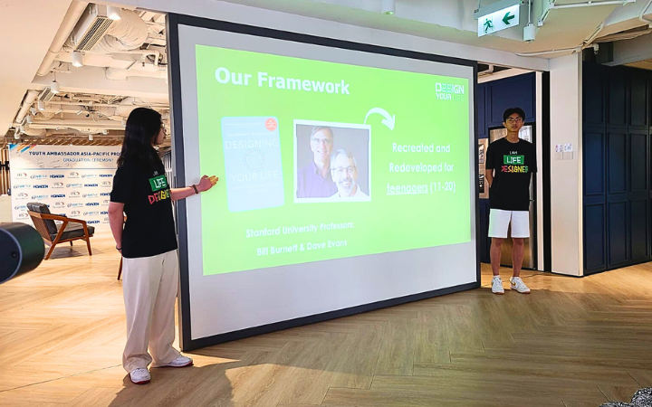 Two teenage individuals stand in front of a presentation screen. They are both facing the audience, with one speaking and gesturing towards the screen.
