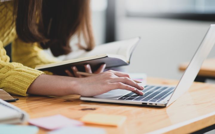 A woman is typing on her laptop and holding her notes.